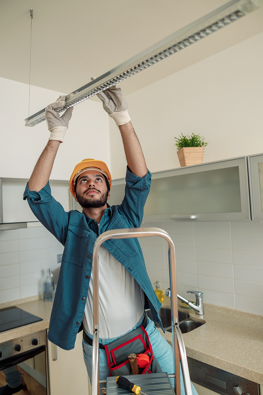 Professional electrician in uniform and hard hat installing a lighting fixture in a kitchen, standing on a ladder with tools in his belt.