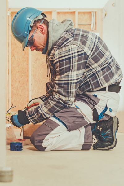 Electrician in a blue hard hat performing maintenance on a wall socket in a construction setting.