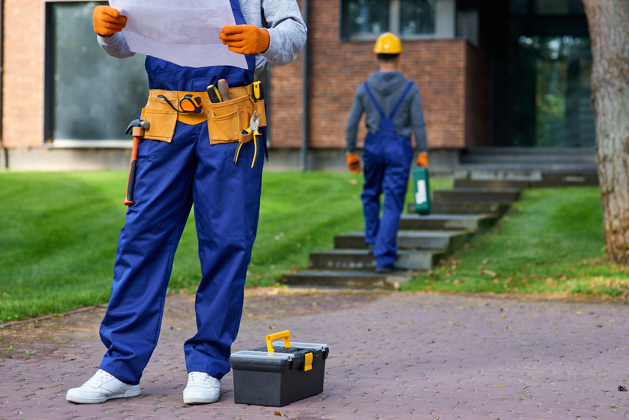 Male electrician in blue overalls holding a blueprint and standing outdoors with a toolbox, while another builder walks towards a house in the background.