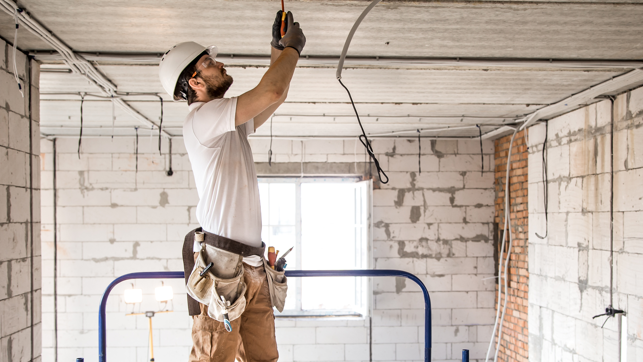 Electrician installing wiring on the ceiling of a construction site, standing on scaffolding with tools in his belt.