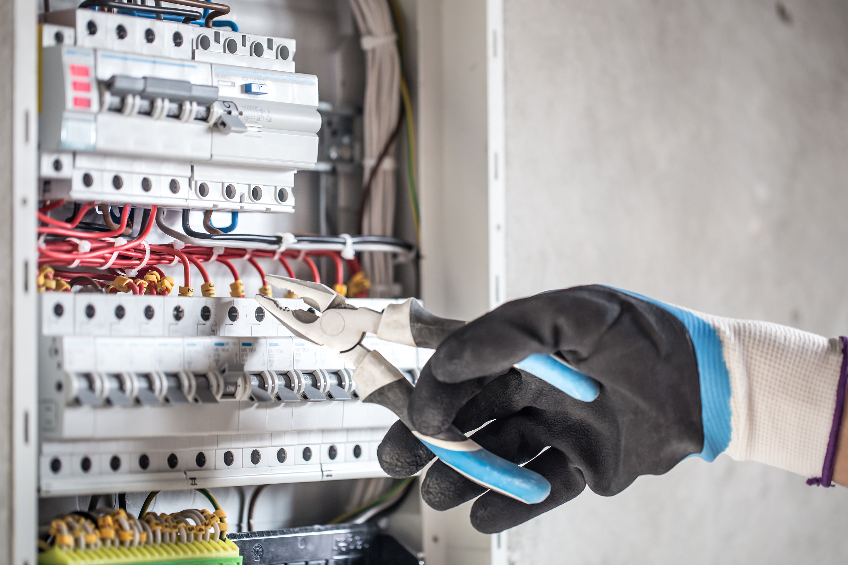 Electrician wearing gloves using pliers to adjust wires in an electrical switchboard, ensuring proper and safe electrical connections.