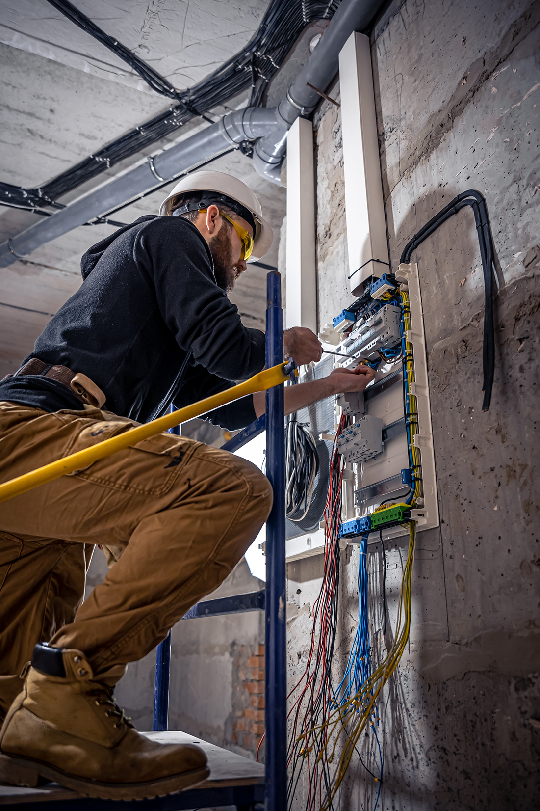 Electrician in safety gear working on a switchboard, organizing wires and ensuring proper electrical connections in a building under construction.