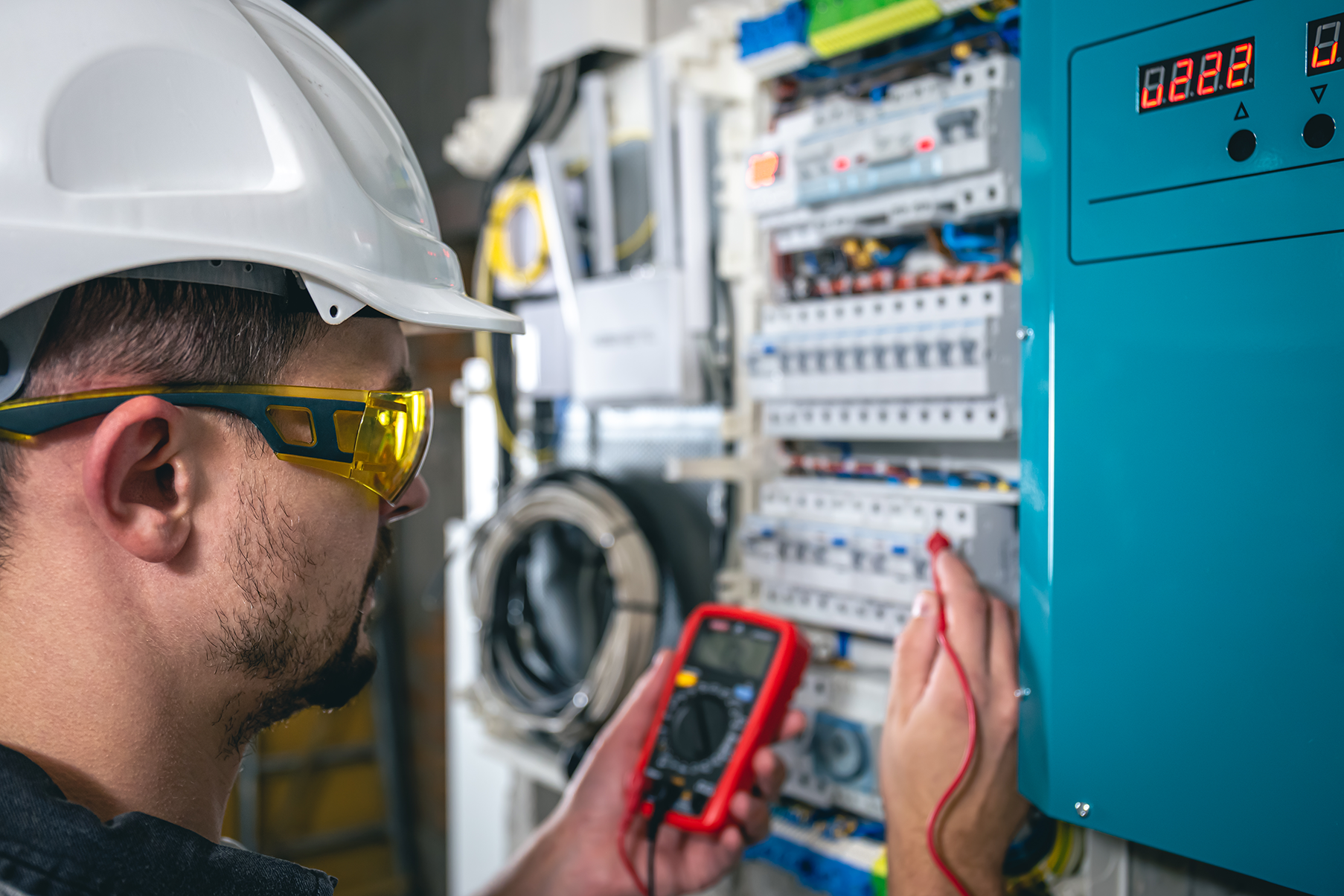 Electrical technician in protective gear using a multimeter to test voltage on a switchboard, ensuring accurate electrical measurements.