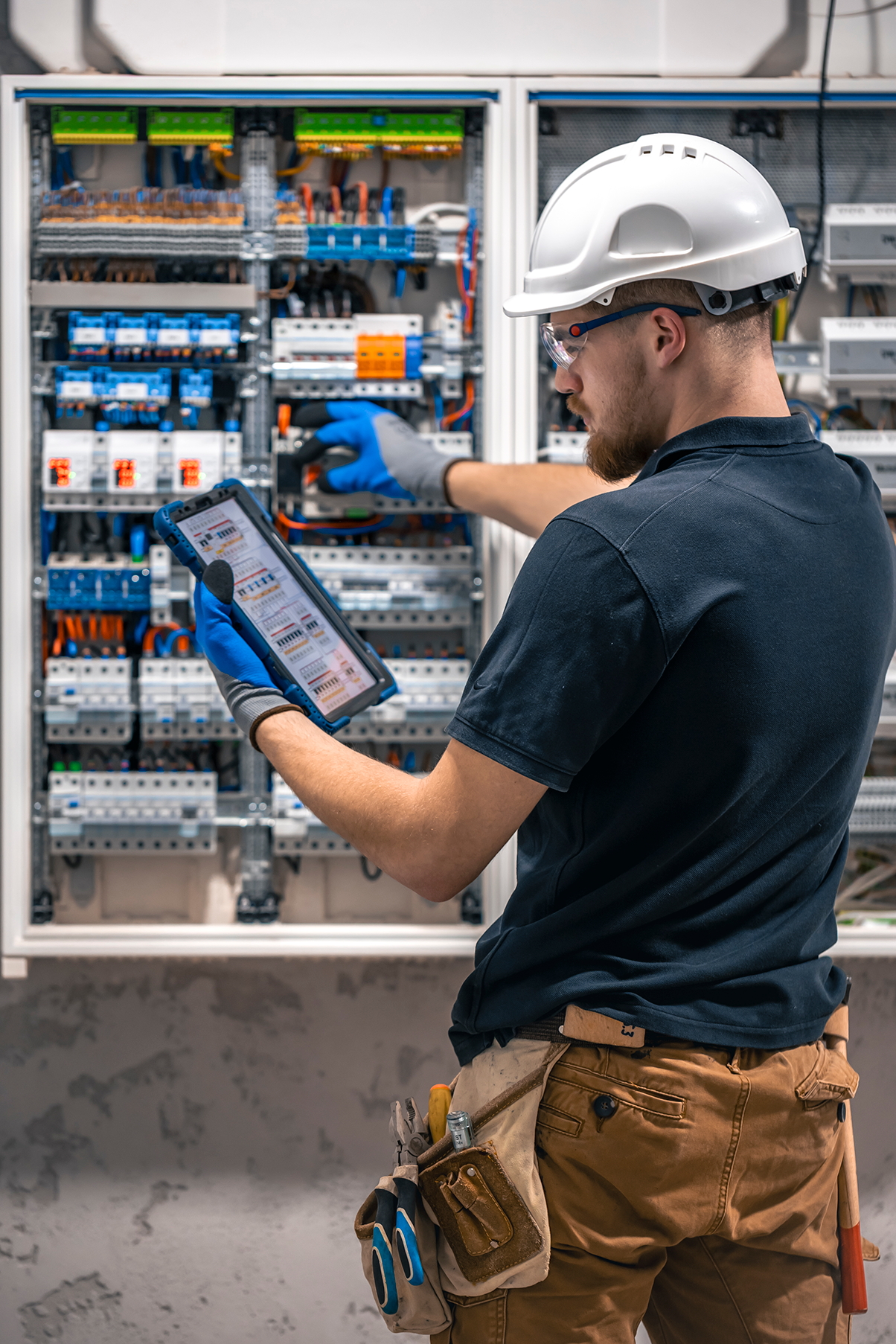 Electrical technician using a tablet to check the switchboard wiring, ensuring accurate and safe connections while wearing protective gear.